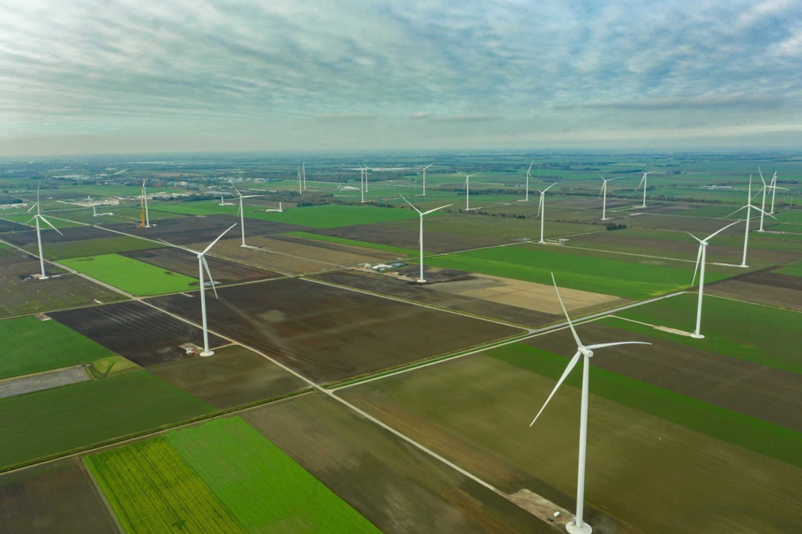 Luchtfoto van een uitgestrekt landschap met talloze windturbines in groene en bruine velden onder een bewolkte lucht.