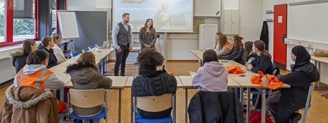 A classroom scene with students seated at tables, listening to two speakers presenting on early careers at OPEA.