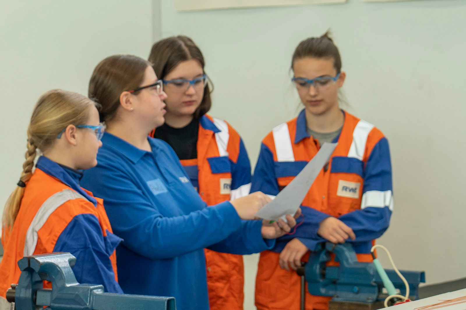 A group of people in safety gear discussing a document, with tools and equipment around them in a workshop.