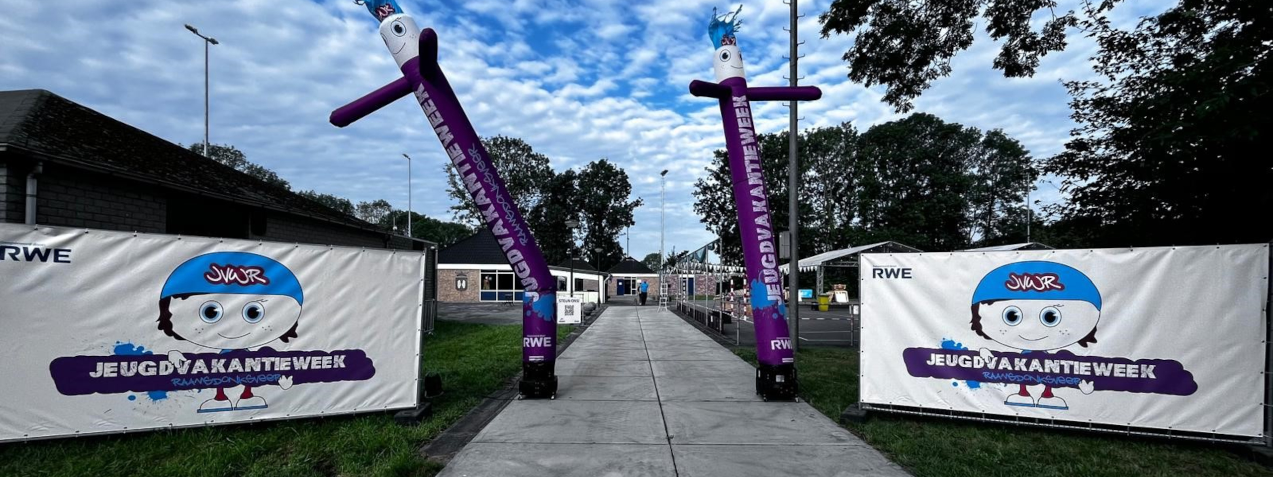 Een kleurrijke ingang versierd met opgeblazen figuren en spandoeken voor een jeugdvakantie-evenement, met een bewolkte lucht.