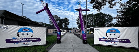 Een kleurrijke ingang versierd met opgeblazen figuren en spandoeken voor een jeugdvakantie-evenement, met een bewolkte lucht.