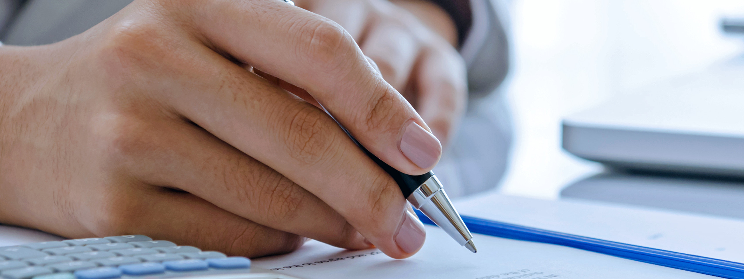 A hand holds a pen above a document, next to a calculator and a laptop.