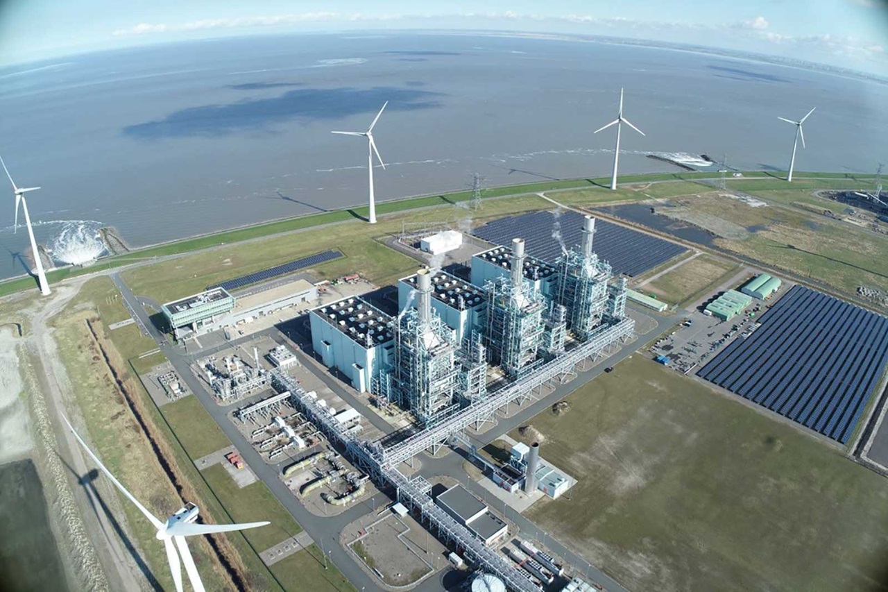 Luchtfoto van een energiepark met een gascentrale, windturbines en zonnepanelen naast het water.
