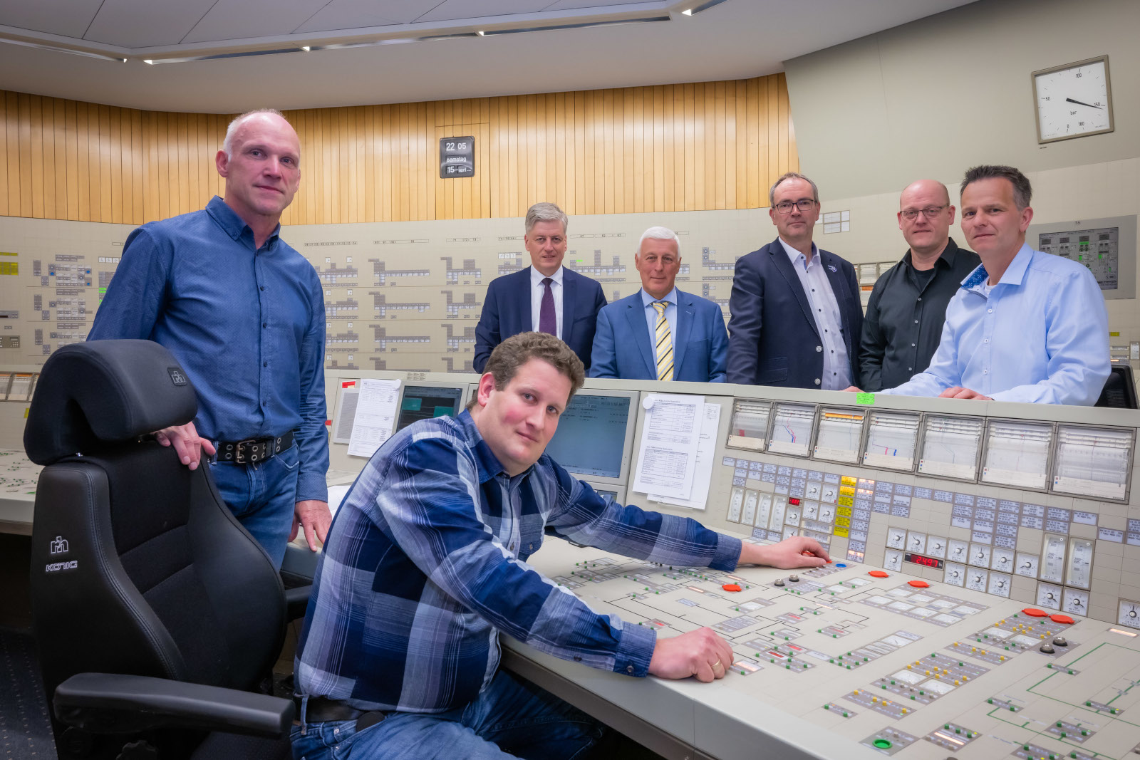 A group of individuals stands by a control panel in a modern room. A man operates the controls.