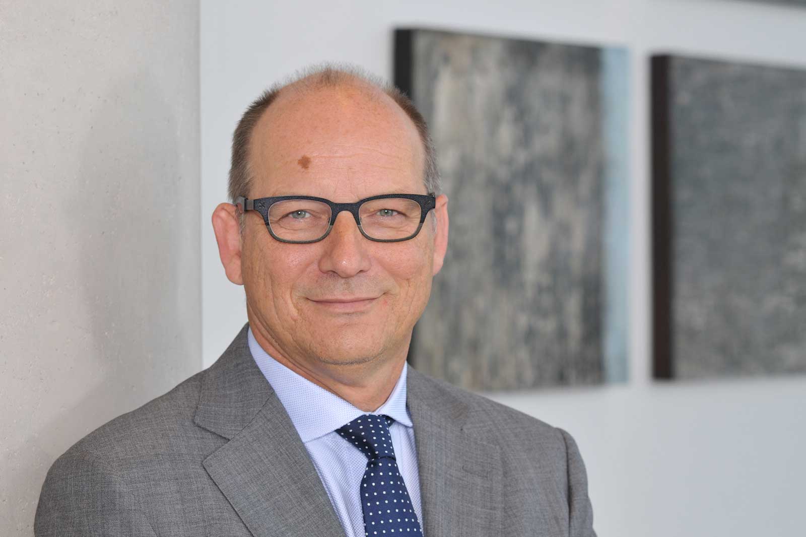 Een man in een pak staat naast een grijze muur met kunstwerken op de achtergrond.