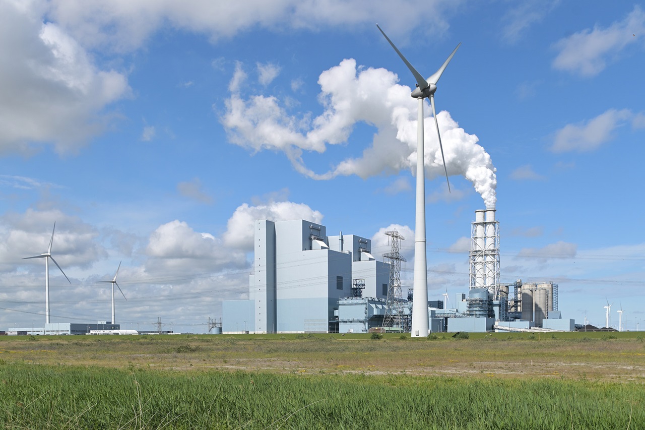 Een industriecomplex met windturbines en rookpluimen tegen een blauwe lucht met wolken.