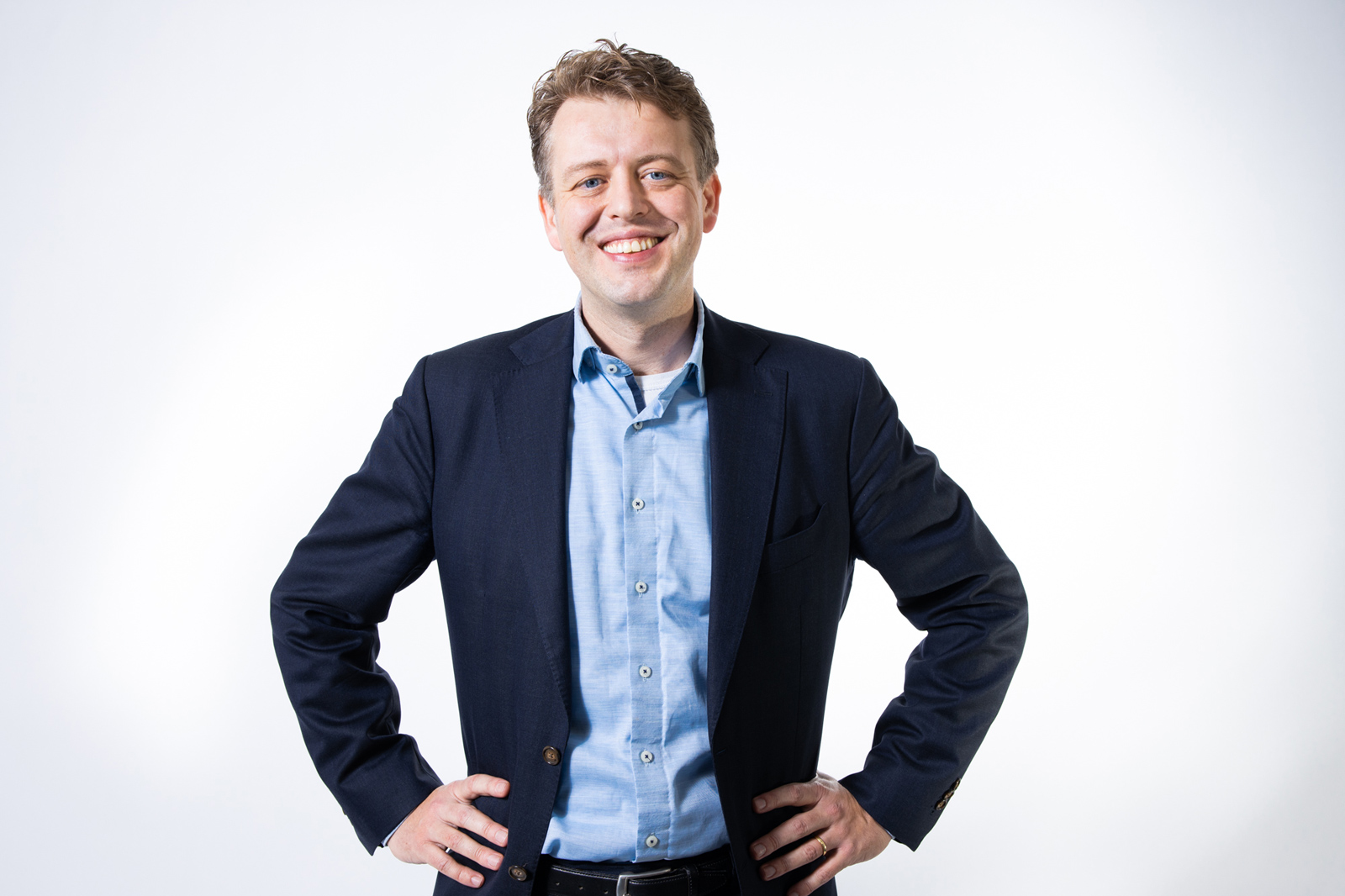 Een man in een blauw shirt en een marineblauwe blazer poseert met handen op de heupen tegen een eenvoudige witte achtergrond.
