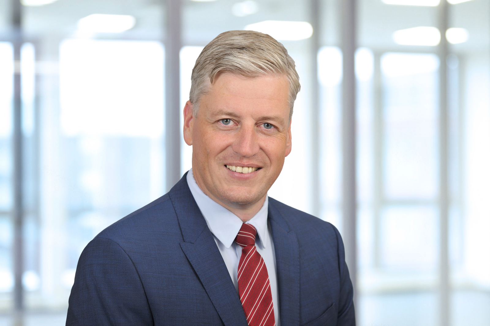 Een man in een donker pak en rode stropdas staat tegen een vervaagde kantoorachtergrond.