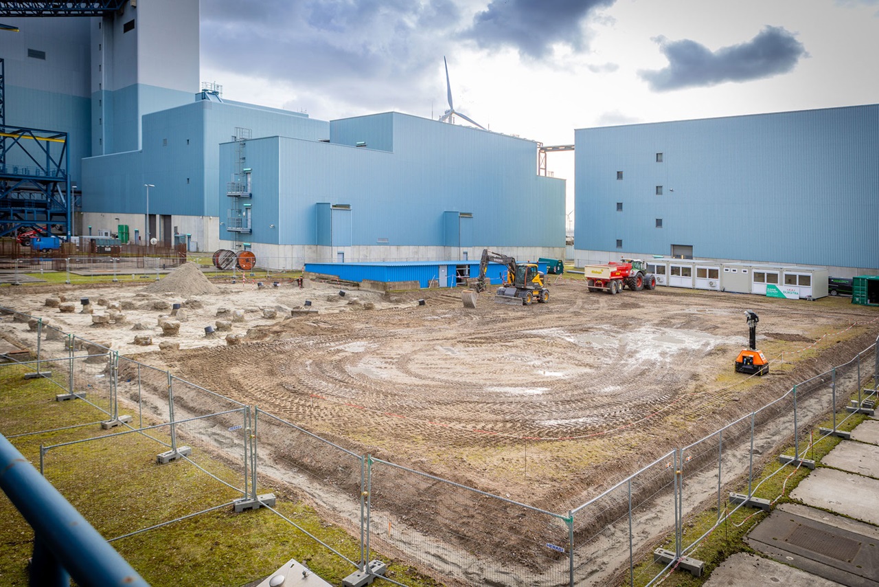 An industrial construction site with machinery and marked ground, surrounded by a fence and buildings in the background.