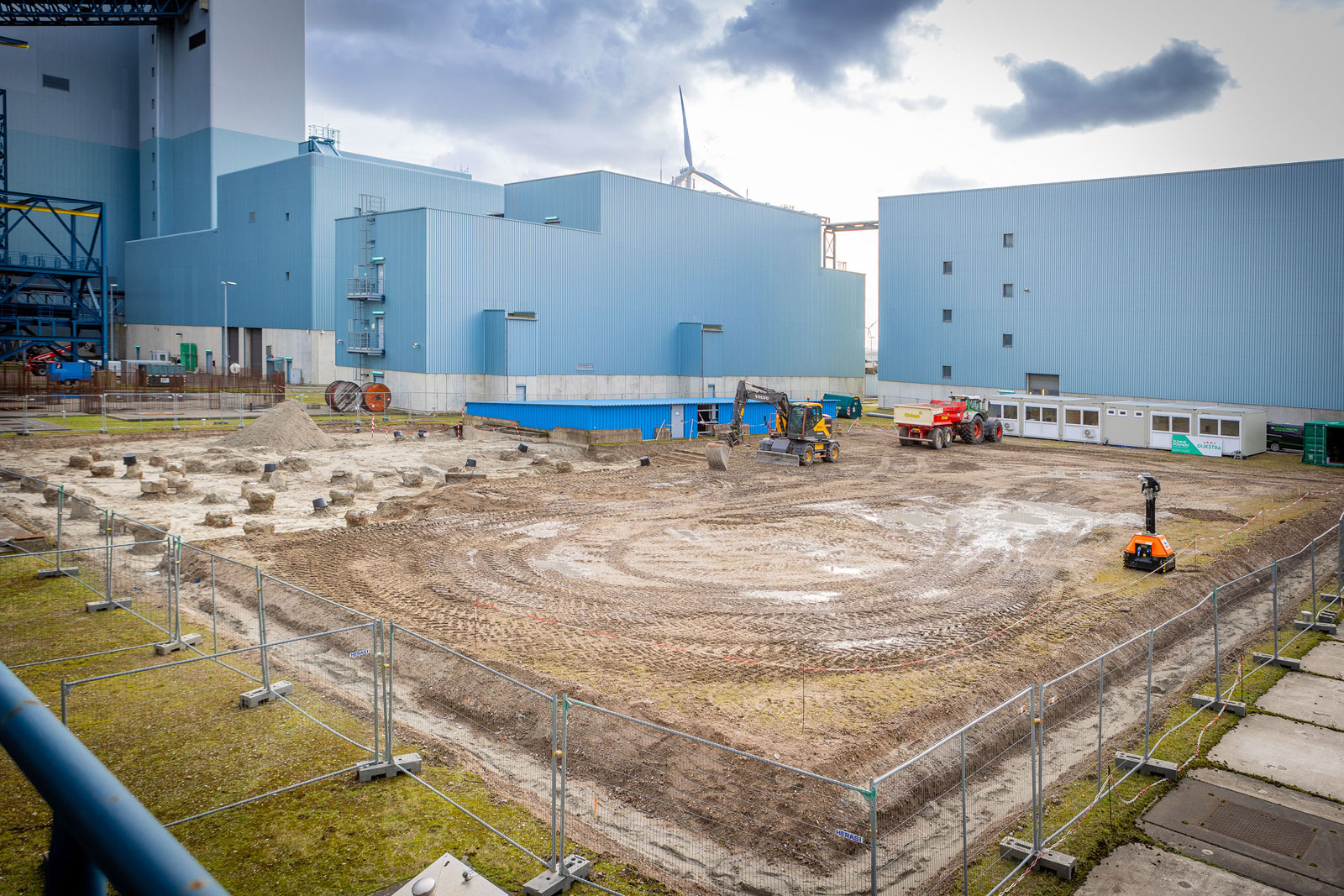 Een industriële bouwplaats met machines en gemarkeerde grond, omringd door een hek en gebouwen op de achtergrond.