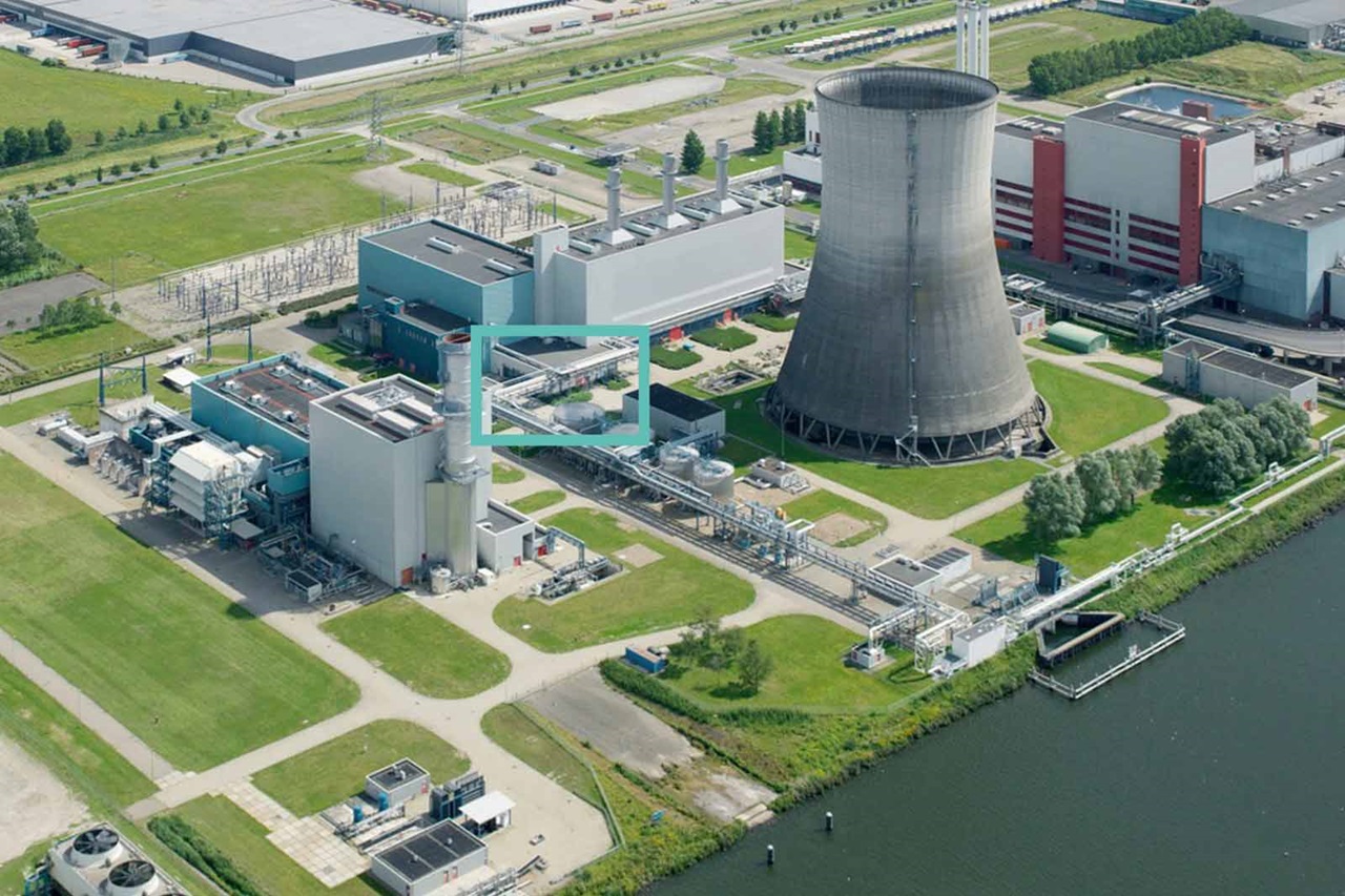 Luchtfoto van een industriecomplex met koeltoren, gebouwen, en elektriciteitsleidingen naast een waterweg.