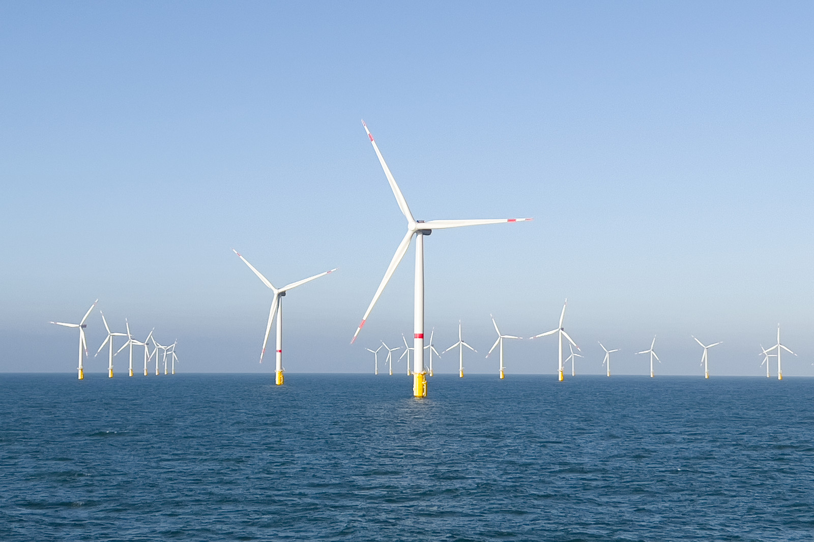 A row of wind turbines stands in the water, under a blue sky, gently reflecting on the calm water surface.