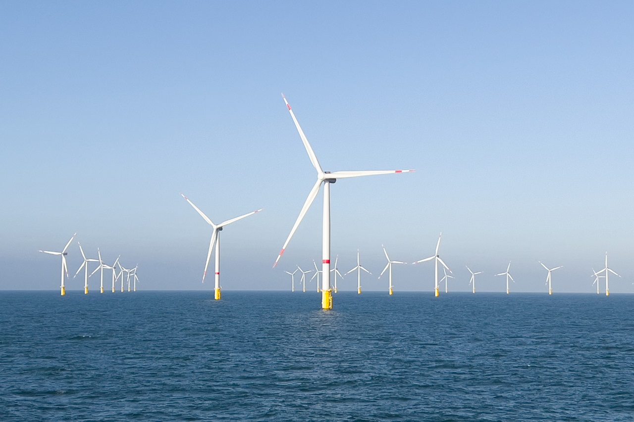 Een rij windturbines staat in het water, onder een blauwe lucht, die zachtjes reflecteert op het rustige wateroppervlak.