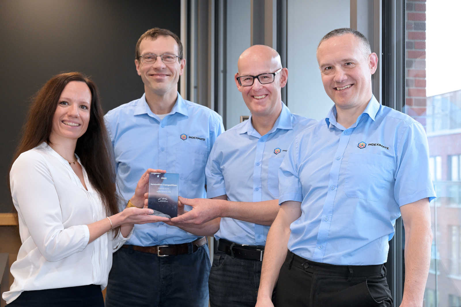A group of four individuals in blue shirts presenting an award. The background features large windows and a modern interior.