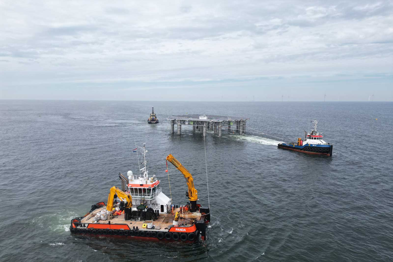 Three vessels manoeuvre near a platform in the ocean, with one boat towing and two others in attendance under a cloudy sky.