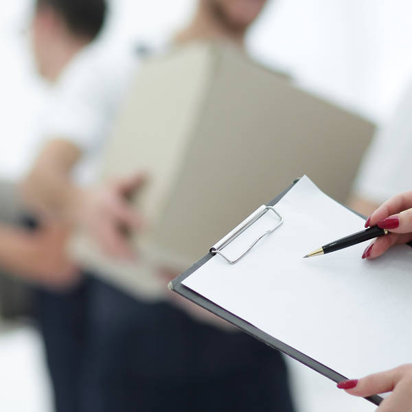 A person holds a clipboard with a pen while another individual carries a box in the background.