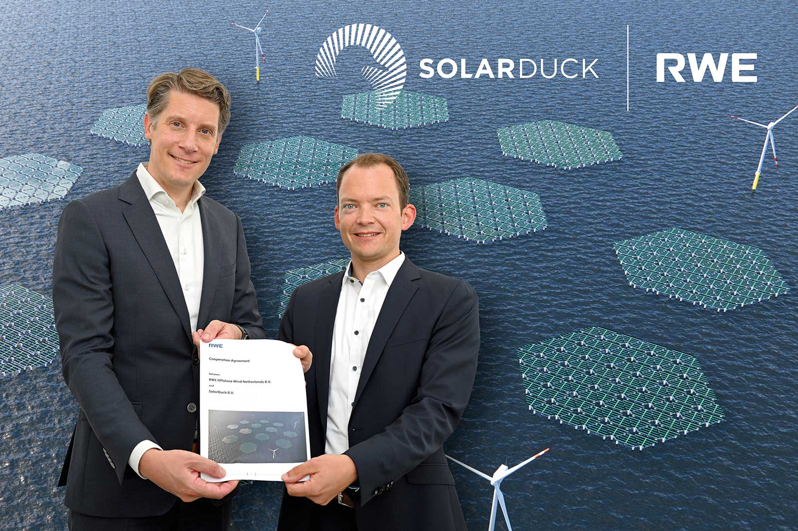 Two individuals hold a cooperation agreement near a backdrop featuring solar panels and wind turbines on water.