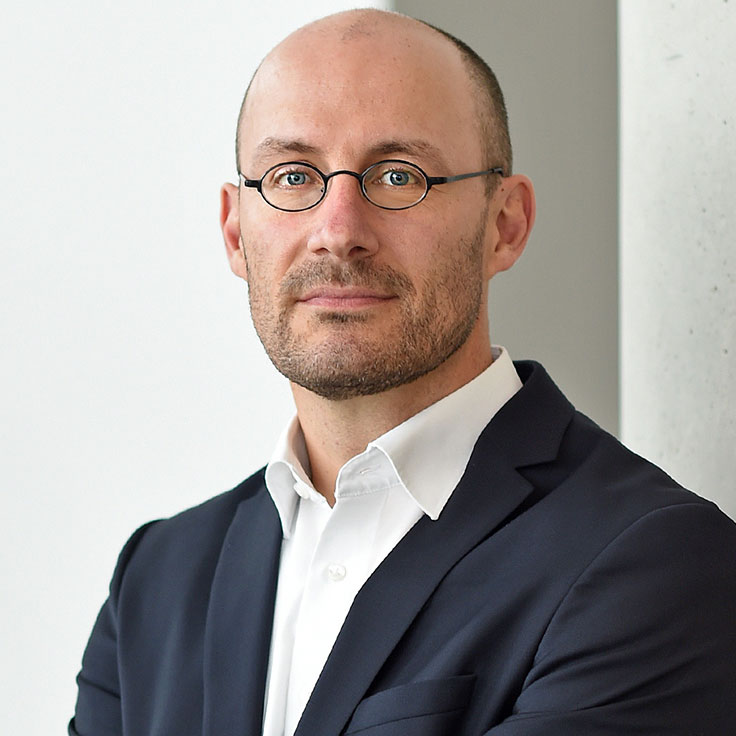 Een man in een donkerblauw pak met een wit shirt staat voor een lichte muur en lijkt professioneel.