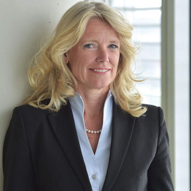 Een vrouw met blond, krullend haar in een zwarte blazer en een parelketting staat voor een raam.