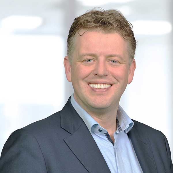 Een man in een blazer en shirt staat tegen een onscherpe achtergrond, wat een professionele sfeer creëert. Portret van Marinus Tabak.