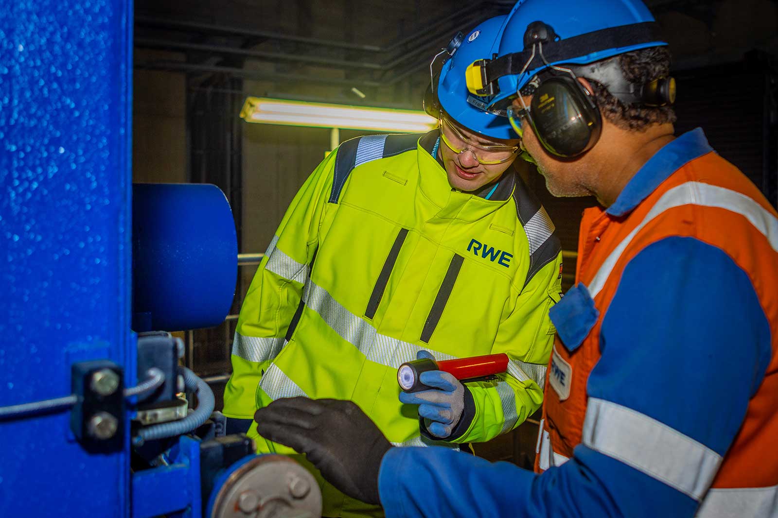 Two engineers in safety gear examine machinery with a flashlight in a dimly lit industrial environment.