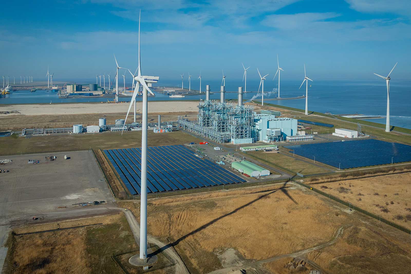 Aerial view of a renewable energy facility, featuring wind turbines, solar panels, and a power plant near the ocean.