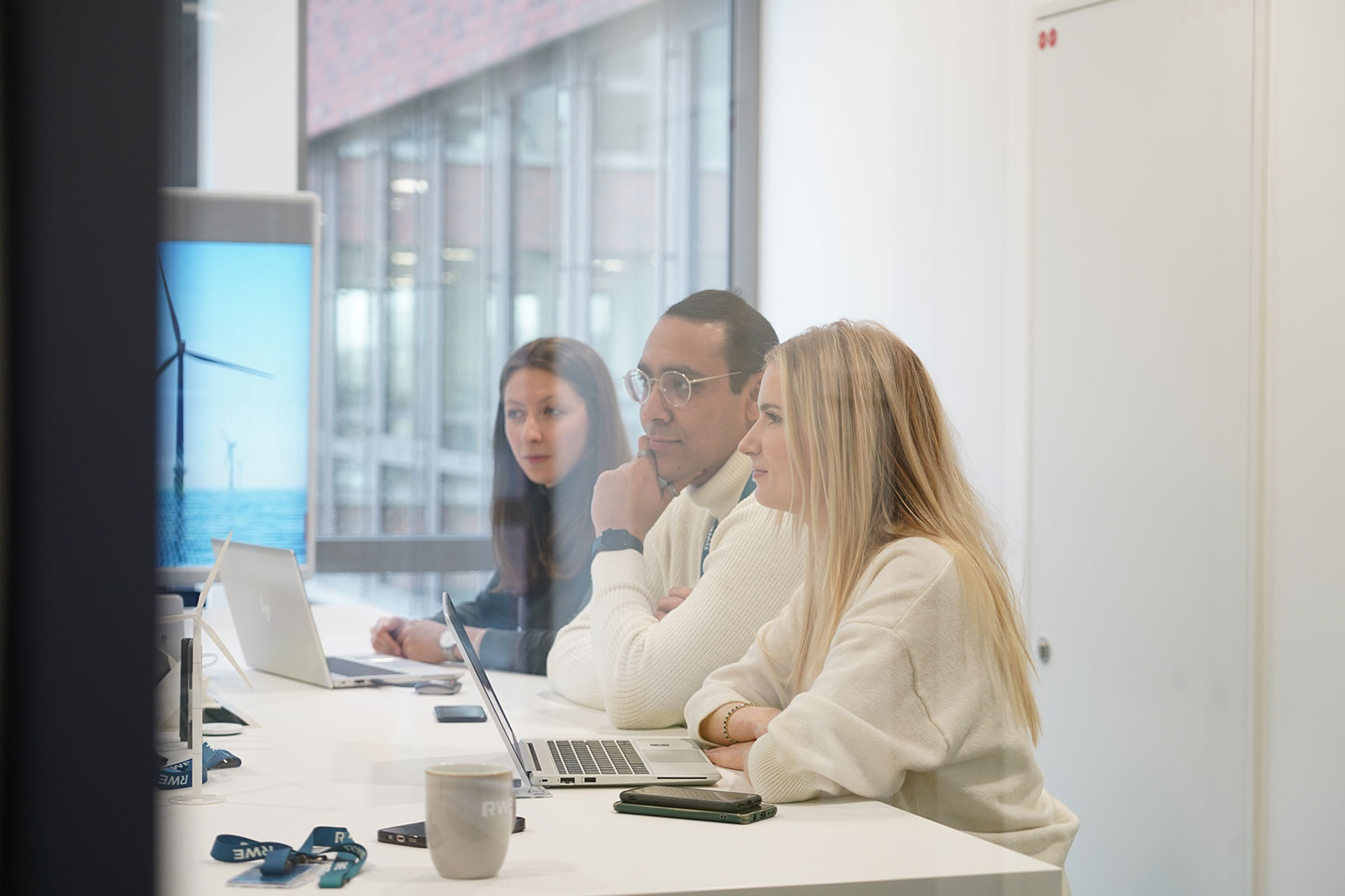 A meeting room with three people engaged in discussion, laptops on the table, and a wind turbine image on the screen.