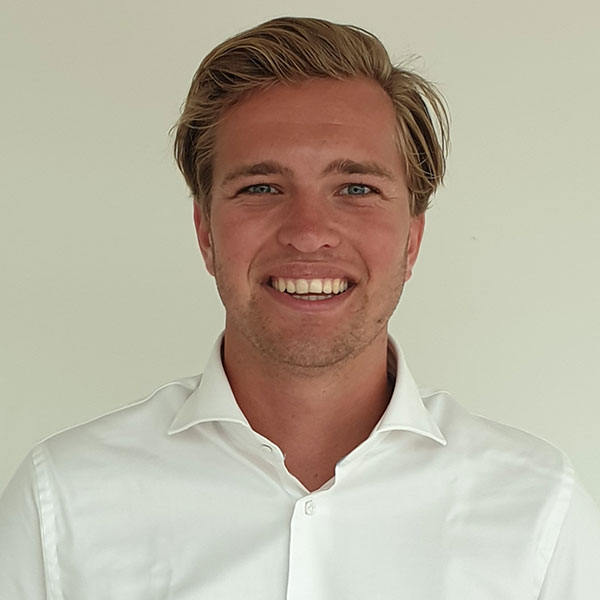 A person in a white shirt with light brown hair stands against a plain background, looking directly at the camera.