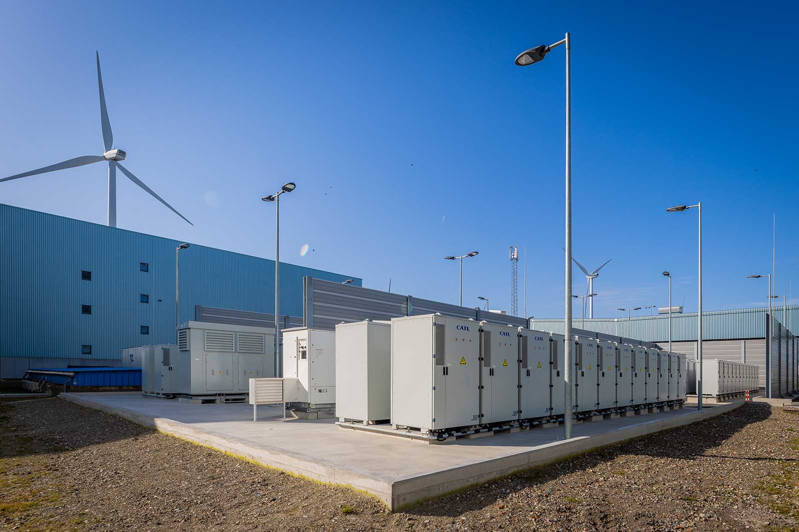 Een zonne-energie-installatie met verschillende grijze kasten, een blauw gebouw, windturbines en straatlantaarns onder een heldere blauwe lucht.