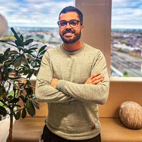 A person stands with arms crossed, wearing a grey sweater, beside a plant and a view of the city in the background.