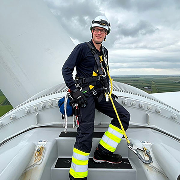 A technician in a safety harness stands on top of a wind turbine, overlooking a cloudy landscape.