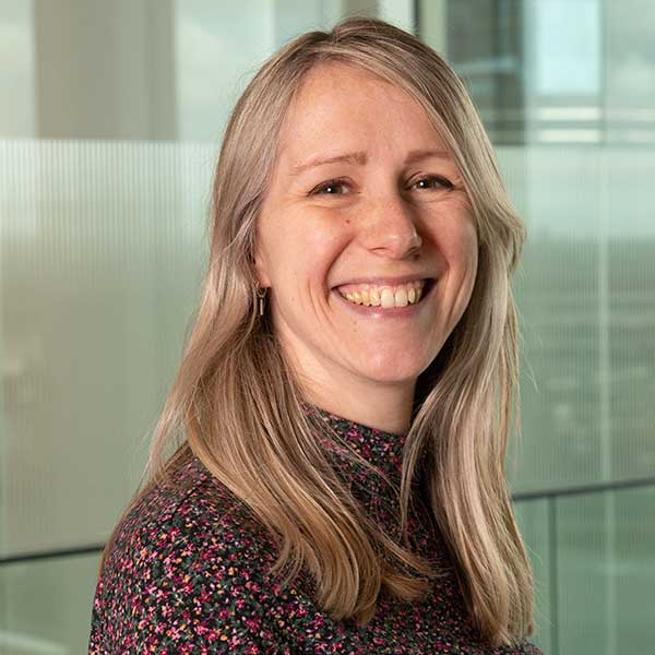 A woman with long blonde hair stands in an office environment, wearing a patterned top, with glass partitions in the background.