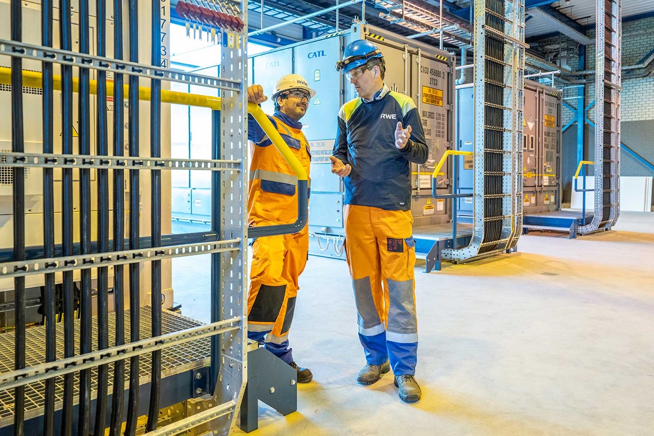 Two workers in orange and blue uniforms converse in a bright industrial facility with storage containers and safety equipment.