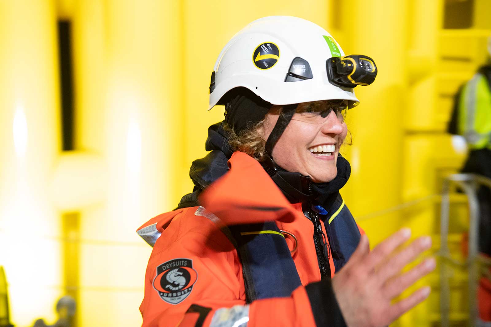Een veiligheidsmedewerker in een oranje drysuit en witte helm gebaart met hun hand in een industriële omgeving met een gele achtergrond.