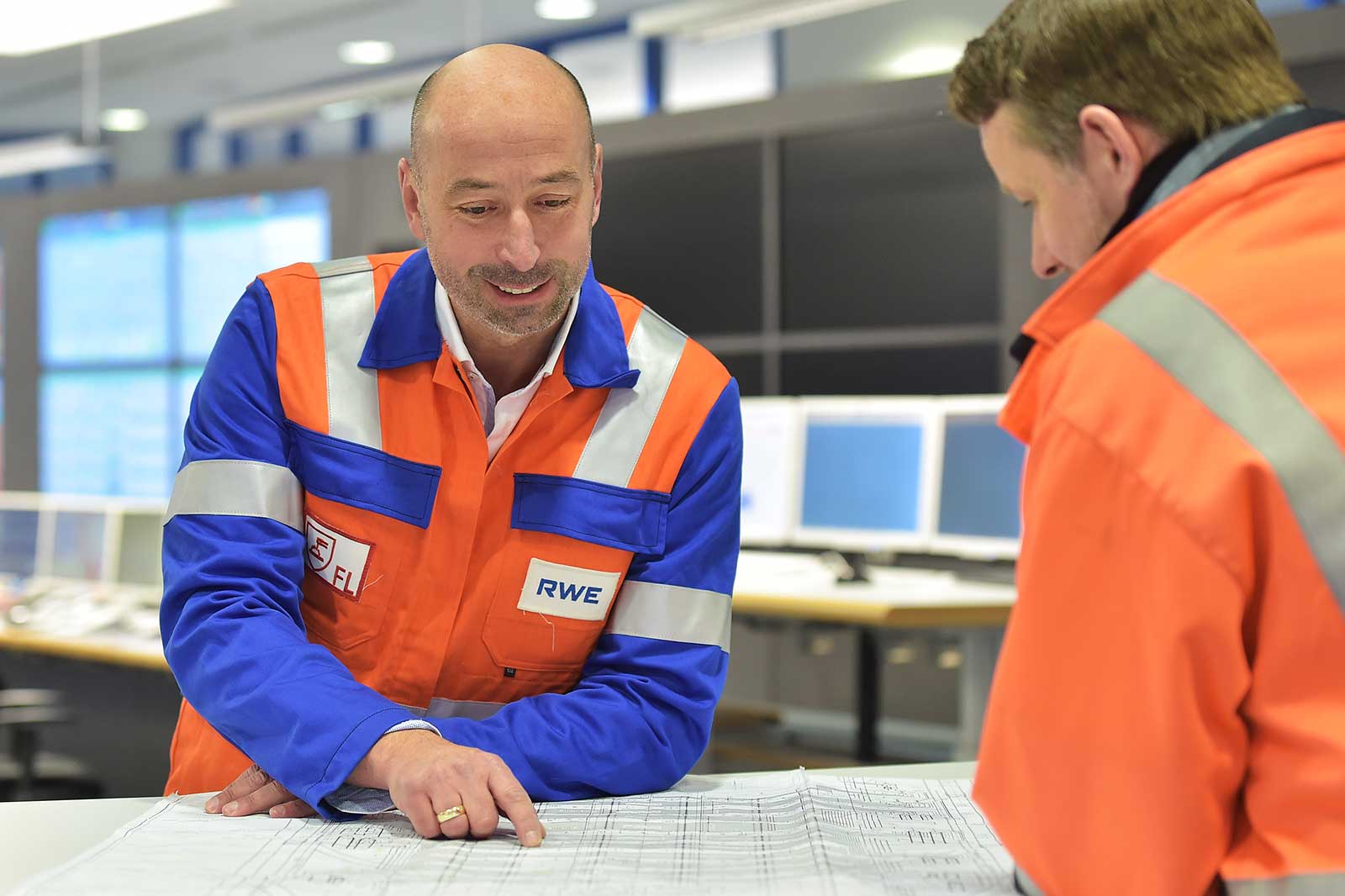 Two workers discuss plans over a blueprint in an industrial setting, wearing orange and blue safety jackets.