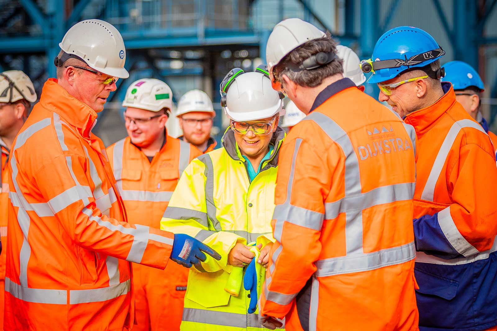 A group of workers in high-visibility orange jackets and hard hats is engaged in discussion at a construction site.