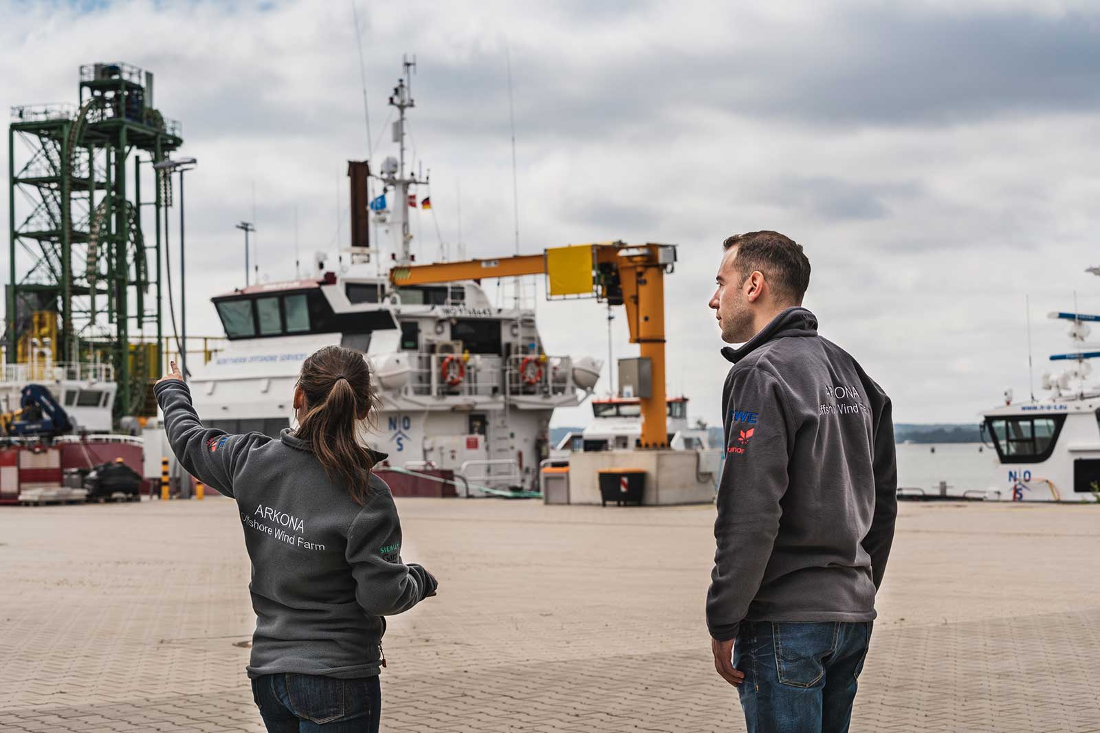 Two individuals stand at a port discussing operations near vessels and cranes, dressed in grey jackets with logos.