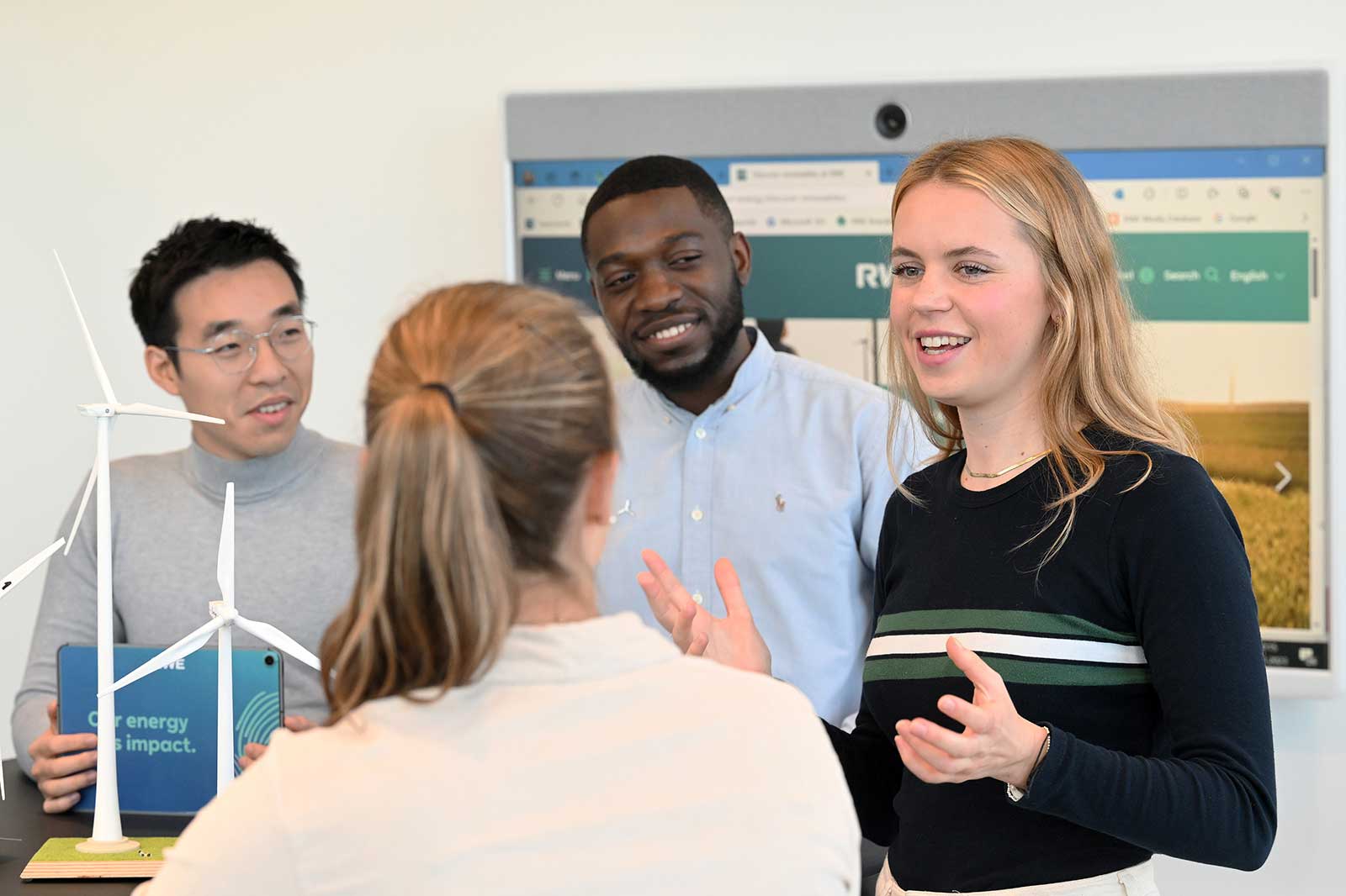 A group of individuals discussing renewable energy with wind turbine models on a table and a digital display in the background.