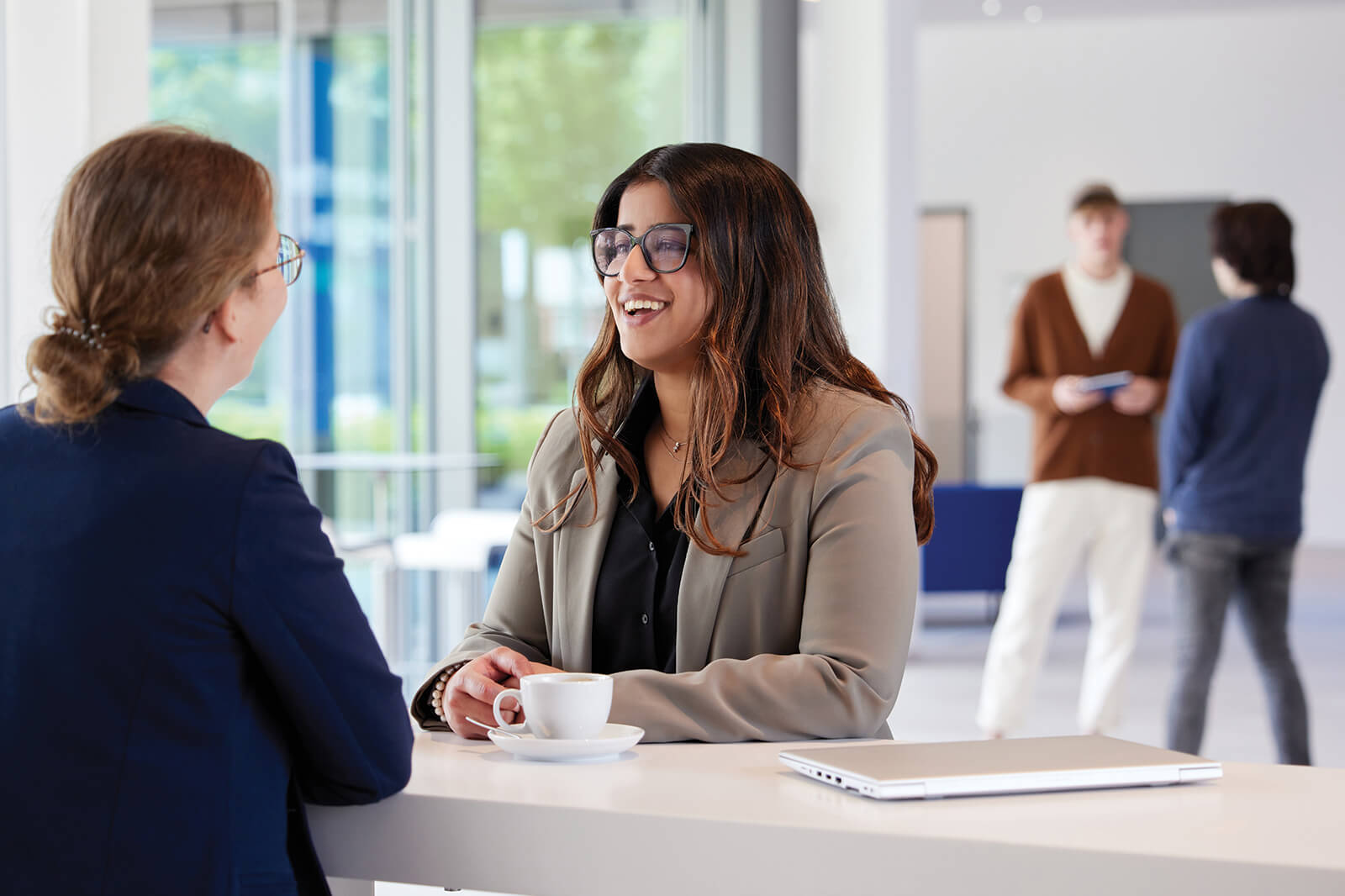 Two women are engaged in conversation at a bright, modern café setting with a laptop and coffee on the table.